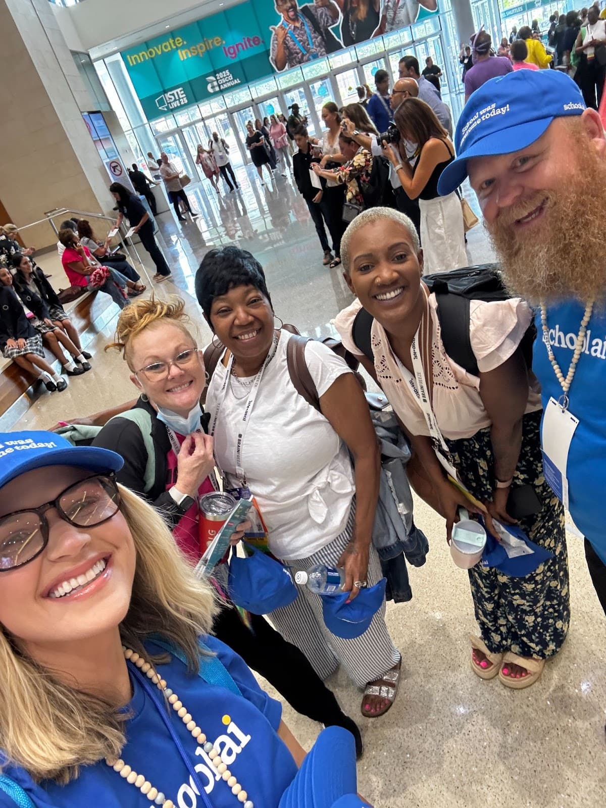 A group of smiling people wearing blue Scholastic branded caps and lanyards pose together at what appears to be a large…