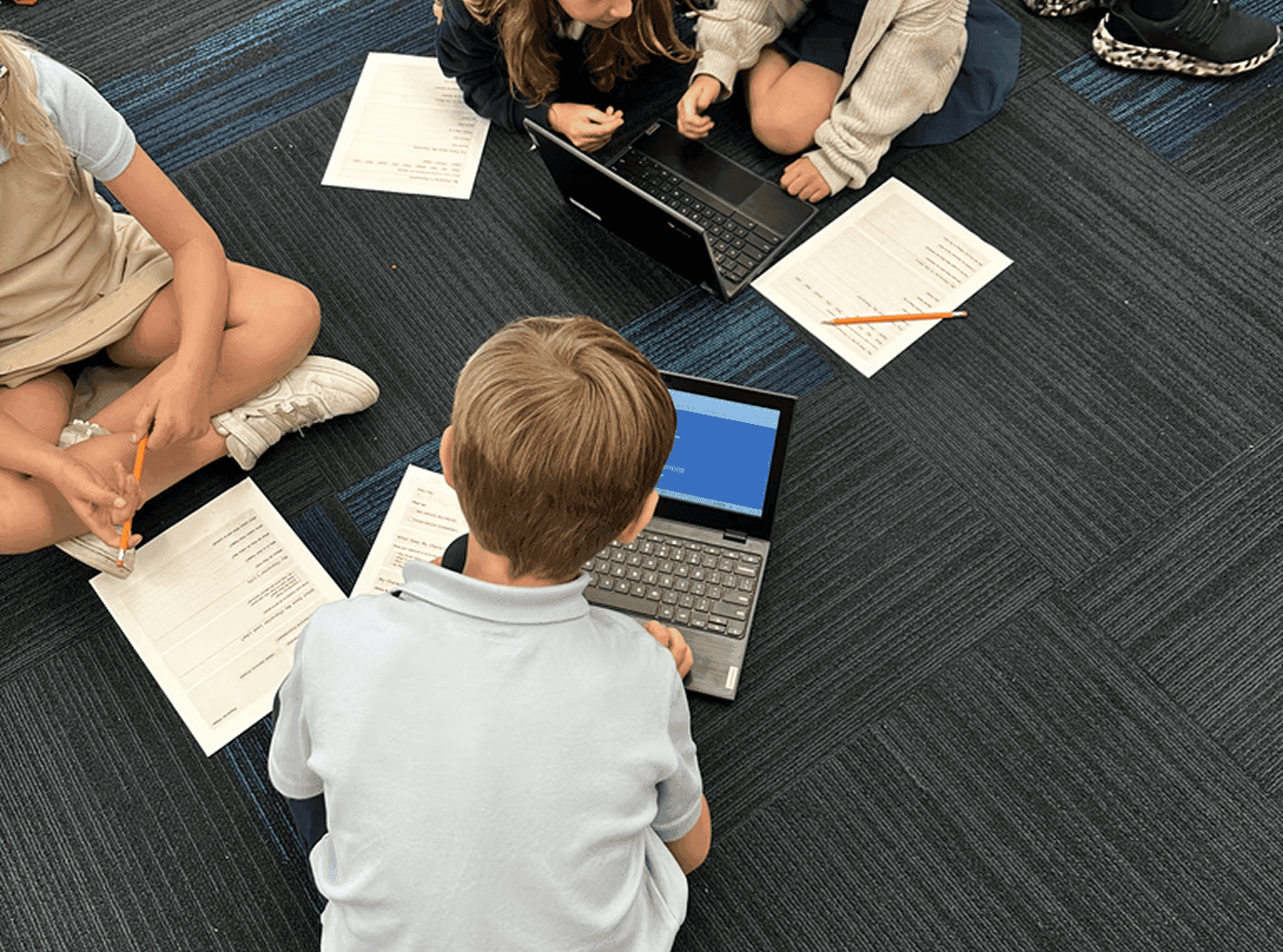 Overhead view of students collaborating with laptops and worksheets on the classroom floor